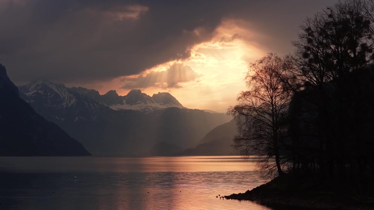 Dramatic sunset light breaks through clouds over a calm lake with silhouetted trees