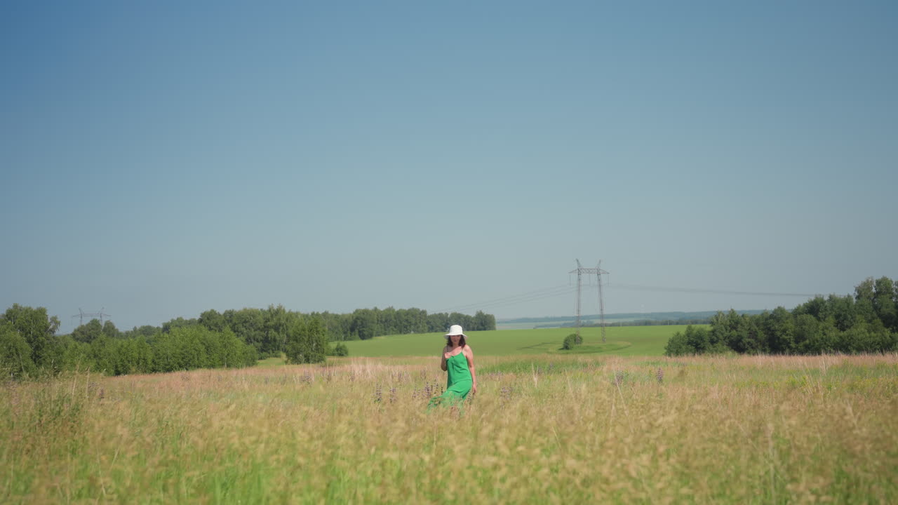 Back view of lady turning in tall swaying grass wearing flowing green summer gown and wide white hat with utility pole and power lines visible beyond lush bright meadow under clear blue sky
