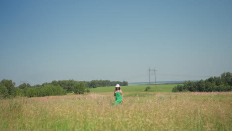 Back view of lady turning in tall swaying grass wearing flowing green summer gown and wide white hat with utility pole and power lines visible beyond lush bright meadow under clear blue sky