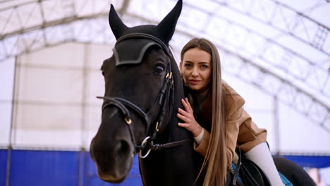 beautiful lady sitting on a black horse leans closer to animal's head. horse riding sport in the drill-hall. low angle view. blurred backdrop.