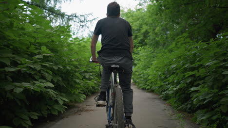 back view of cyclist cycling along a paved path surrounded by lush greenery, white road markings guide the way as the cyclist rides, with a blurred view of another cyclist ahead in the distance