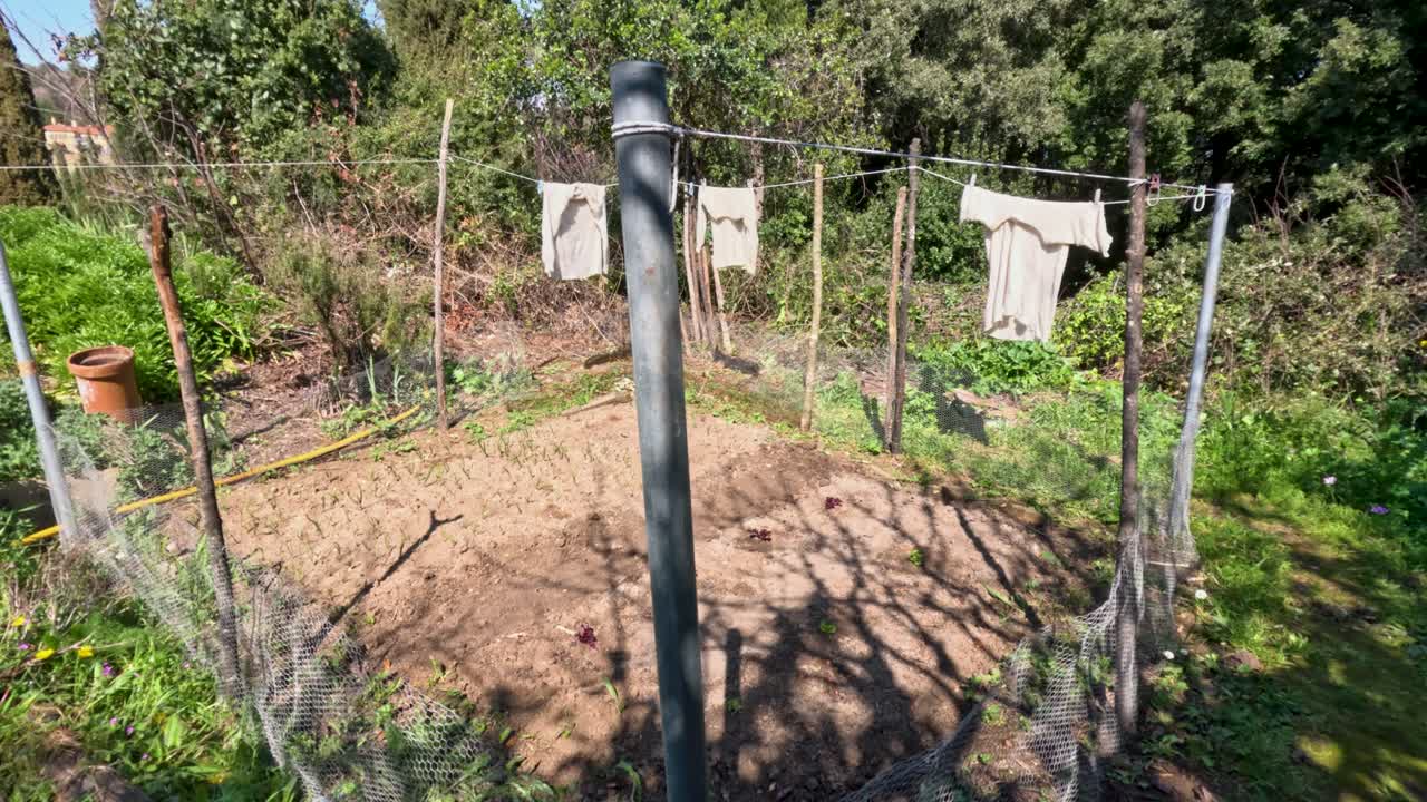 Clothes are hanging on a line in a garden area. The ground is bare and has rows marked for planting. Greenery and trees surround the space