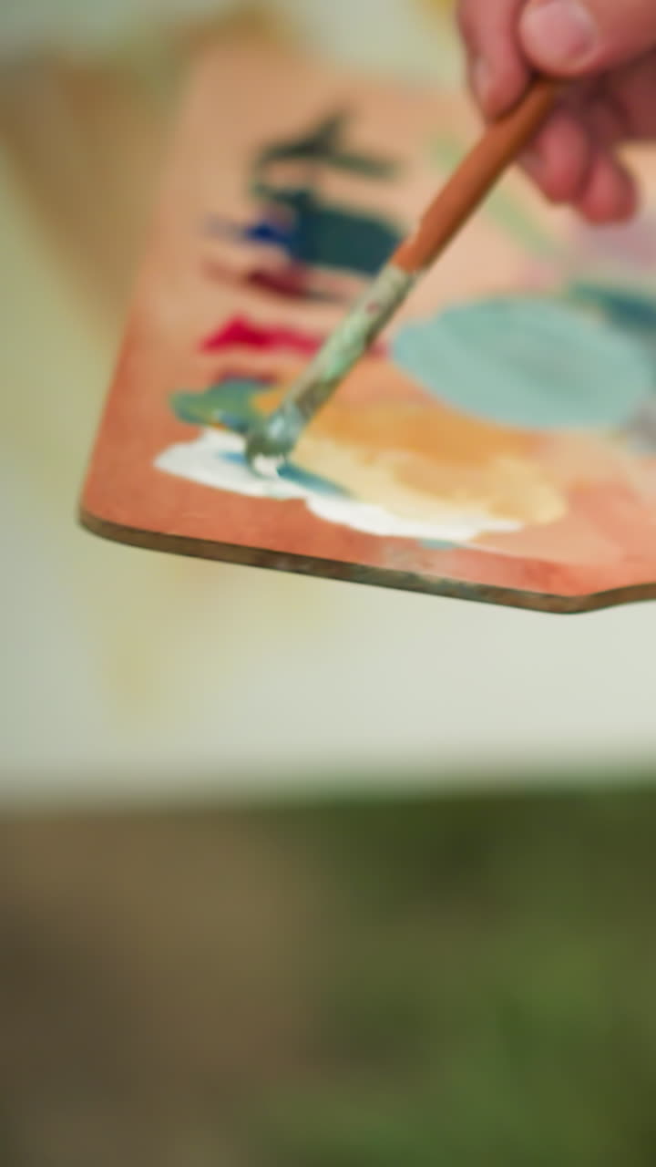 A painter delicately holds a wooden palette filled with vibrant paint as they apply final touches to their artwork. focusing on the hand and brush in action