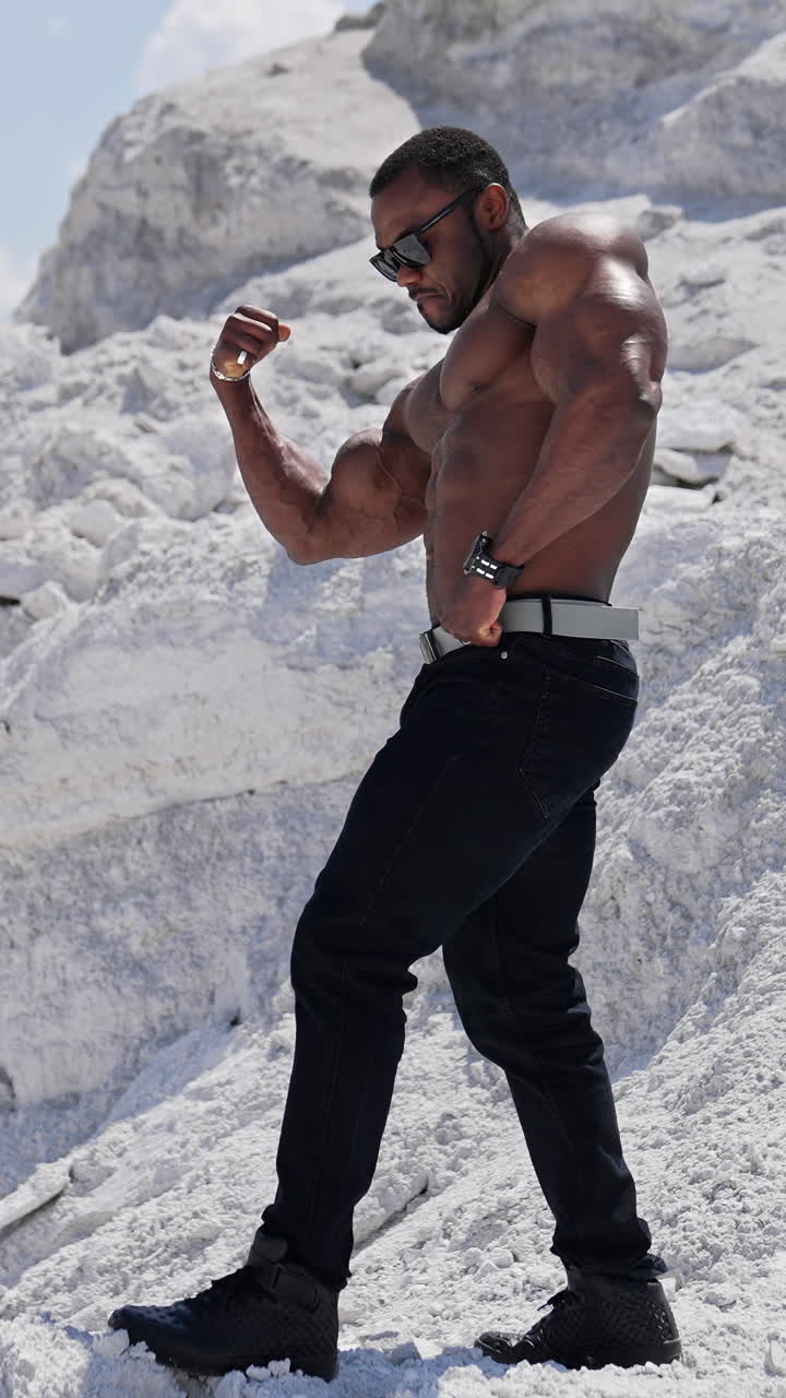 Muscular athlete on white rocky background. African american man in black glasses without shirt showing his biceps. Profile view. Vertical video