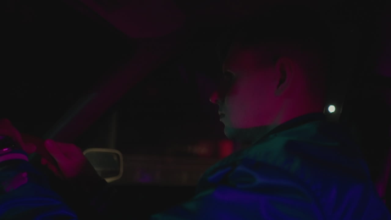 Interni auto di notte, uomo alla guida con le mani salde sul volante e sguardo concentrato, riflessi di insegne al neon e lampioni sul vetro, guida tranquilla lungo il viale urbano, profilo pensieroso, ritmo lento, skyline in lontananza