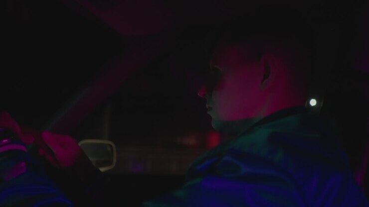 Interni auto di notte, uomo alla guida con le mani salde sul volante e sguardo concentrato, riflessi di insegne al neon e lampioni sul vetro, guida tranquilla lungo il viale urbano, profilo pensieroso, ritmo lento, skyline in lontananza