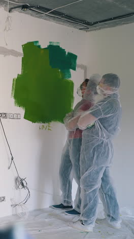 dolly cam shot: funny young family in protective workwear and masks comes up dancing to large green spot on white wall in room
