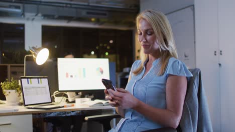 video de una mujer caucásica usando un teléfono inteligente, trabajando hasta tarde en la oficina