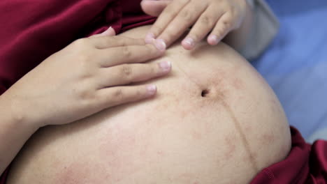 an expectant woman who is lying on her back is stroking her swollen and bare pregnant belly with her fingertips