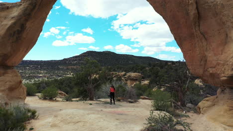 Aerial-View-of-Young-Female-Photographer-Standing-Under-Amazing-Natural-Arch-in-Desert-Landscape-of-New-Mexico,-Navajo-Nation-Territory,-Drone-Shot