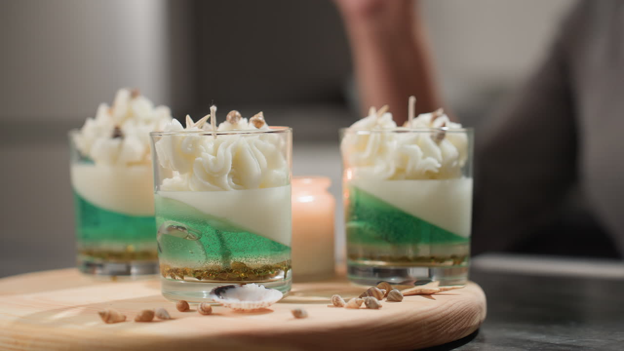 Three beautifully decorated candle jars with whipped cream design placed on wooden tray surrounded by small seashells while blurred hand of joyful chandler grooves and gestures happily in background