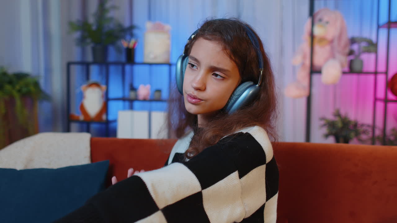 Happy young girl in wireless headphones relaxing sitting on sofa at home listening favorite music