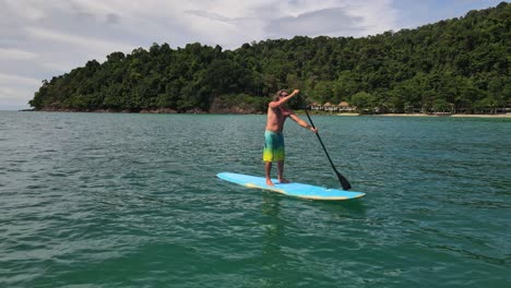 Niedriger-Winkel-Nach-Hinten-Dolly-Shot-Drohnenansicht-Eines-Kaukasischen-Mannes,-Der-Auf-Einem-Sup-Paddelbrett-In-Türkisfarbenen-Tropischen-Klaren-Gewässern-Mit-Strand-Und-Küste-In-Thailand-Trainiert