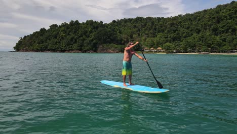 ángulo bajo, tiro de muñeca hacia atrás, vista de dron de un hombre caucásico haciendo ejercicio en una tabla de remo en aguas turquesas tropicales claras, con playa y costa en tailandia
