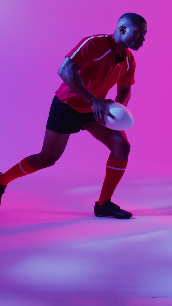Vertical video of african american male rugby player crouching with rugby ball over pink lighting