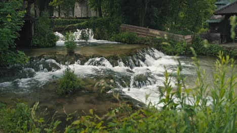 Small-cascading-waterfalls-flowing-through-a-lush-garden-with-stone-buildings-in-Rastoke,-Croatia