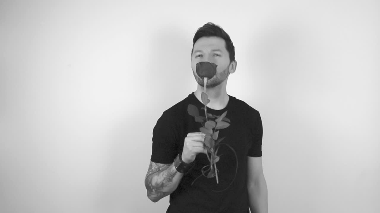 Grayscale footage of a man with tattoos looking and smelling a rose by putting it against his nose and then offering it in front of him with a smile or to the viewer in slow motion. Rose is blossoming