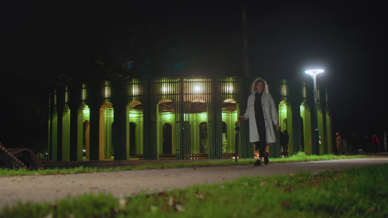 Girl in white coat walking peacefully with umbrella in hand through serene night park, passing under bright streetlights with glowing modern green pavilion structure and people visible in background