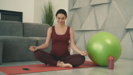 Joven-Mujer-Bastante-Embarazada-Haciendo-Yoga-En-Posición-De-Loto-Y-Meditando,-Usando-Ropa-Deportiva-En-Casa