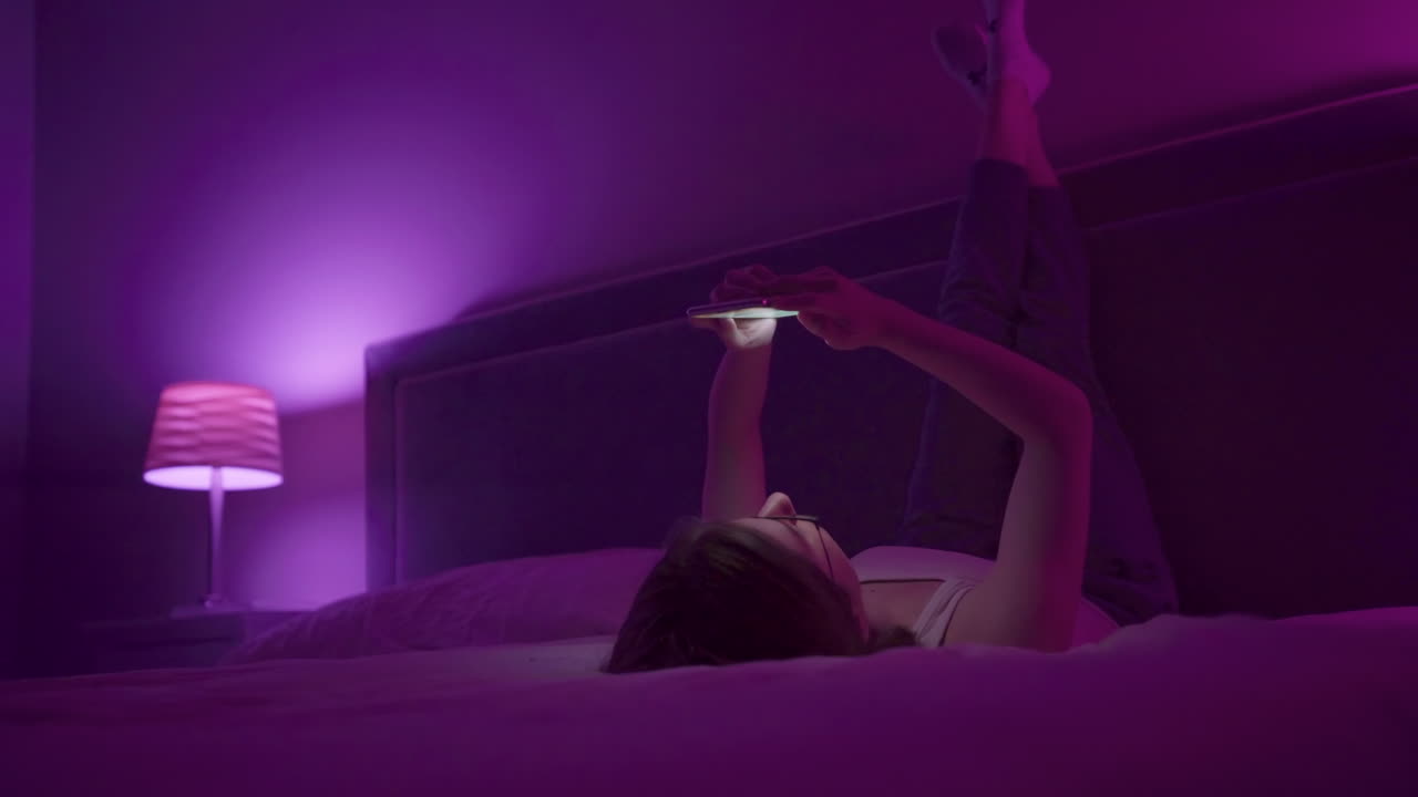 Girl laying on back browses social media on smart phone late at night in bedroom.