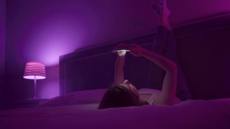 girl laying on back browses social media on smart phone late at night in bedroom.
