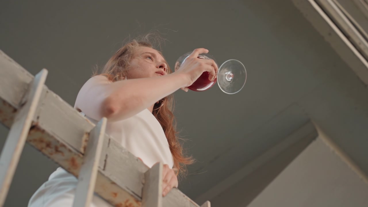 Woman in white dress stands on metal stairs holding and drinking from glass of red liquid. scene features industrial elements like rusted steel beams against soft lighting with subject hair