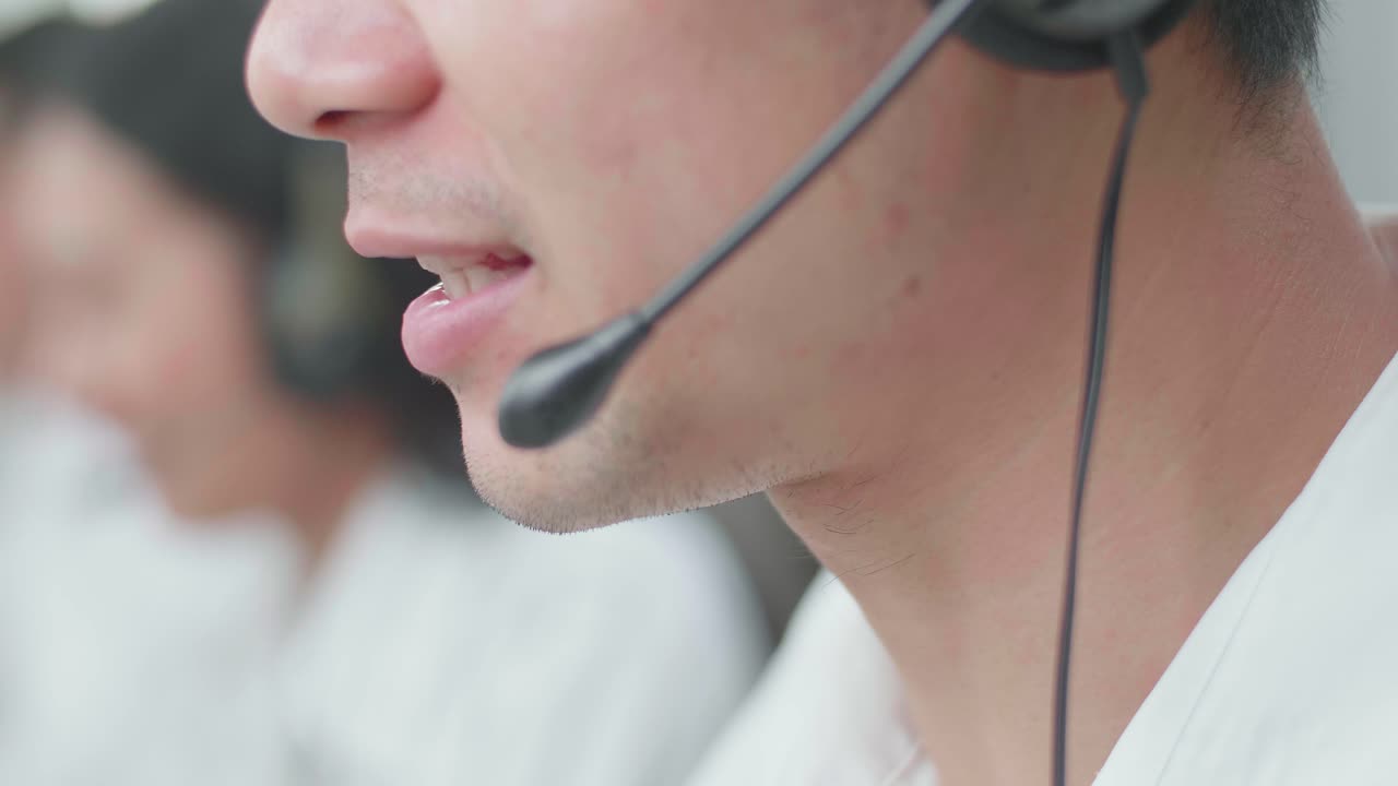 Close Up Of An Asian Man Call Center Agent Wearing Headset Smiling While Speaking To Customer On The Call During Working With His Colleagues At The Office