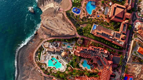 top down aerial drone shot over a beautiful beach resort in tenerife, canary islands, spain. high view of the premises, outdoor pools, the ocean and the volcanic beach. summer holidays destination.