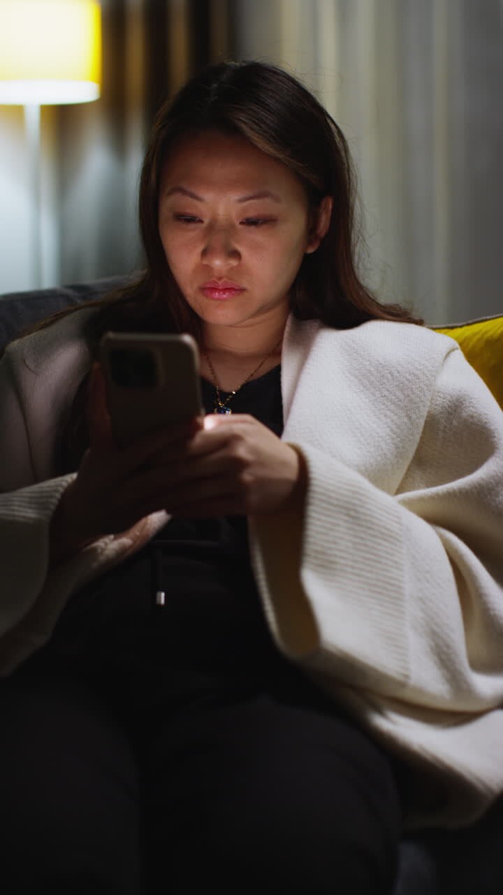 Vertical Video Of Woman Spending Evening At Home Sitting On Sofa With Mobile Phone Scrolling Through Internet Or Social Media 2