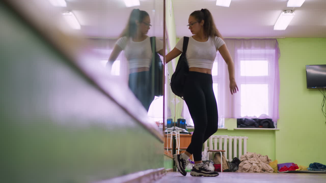 View of girl in casual white top and black leggings carrying school bag walking from door toward window, reflection showing on mirror inside bright indoor space with wooden floor and purple curtains