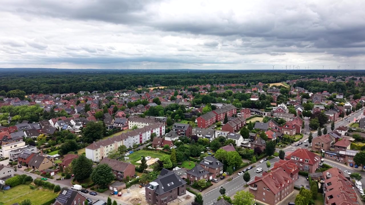 Gęsto zabudowane niemieckie miasto rozciąga się pod ciężkim zachmurzeniem, wysokie apartamentowce i historyczne domy szczytowe skupiają się wzdłuż wysadzanych drzewami ulic pośród czerwonych dachów poniżej. Szerokie ujęcie z drona