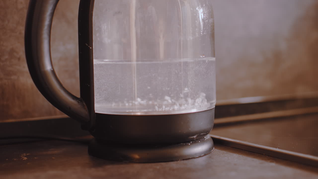 Close up of electric kettle with transparent body boiling water as bubbles rise and steam forms inside glass container placed on kitchen counter showing daily beverage preparation process indoors