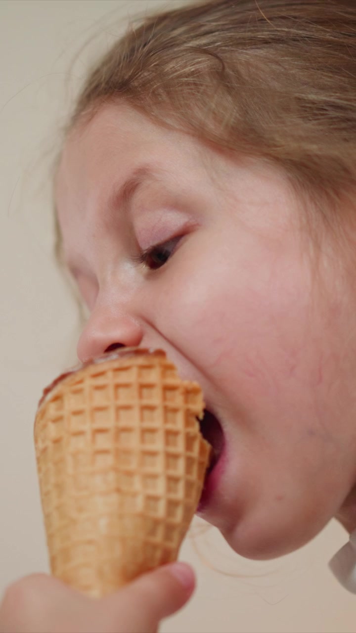 Close up of young girl enjoying ice cream cone repeatedly bringing it to her mouth. waffle texture and soft lighting are visible throughout sequence. Background is plain and light colored