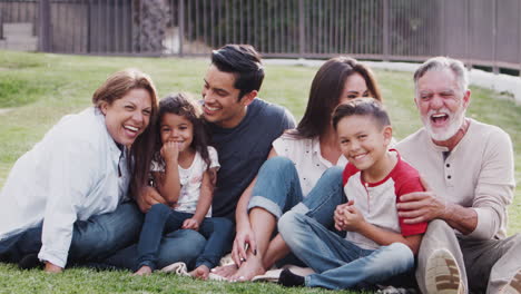 Familia-De-Tres-Generaciones-Sentada-En-El-Césped-Del-Parque,-Haciéndose-Cosquillas-Y-Sonriendo-A-La-Cámara