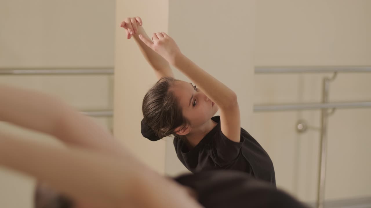 A group of young ballet students in black dancewear practicing positions in a spacious ballet studio with wooden flooring and wall-mounted barres. Focused expressions and synchronized movements.