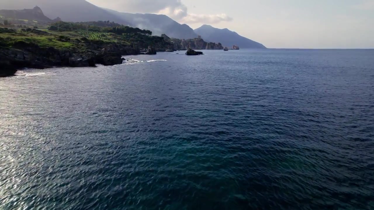 Piccole imbarcazioni riposano vicino a un promontorio roccioso mentre la foschia mattutina si estende sulle verdi colline della costa settentrionale della Sicilia
