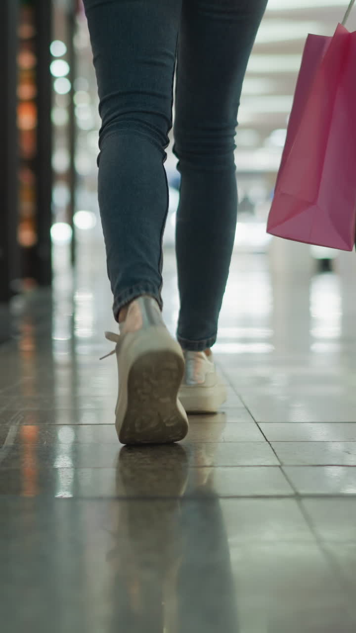 vista de la pierna de una mujer en vaqueros y zapatillas de deporte que lleva bolsas de compras de menta y rosa mientras camina a través de un centro comercial con pisos de azulejos brillantes y reflejos de luz coloridos