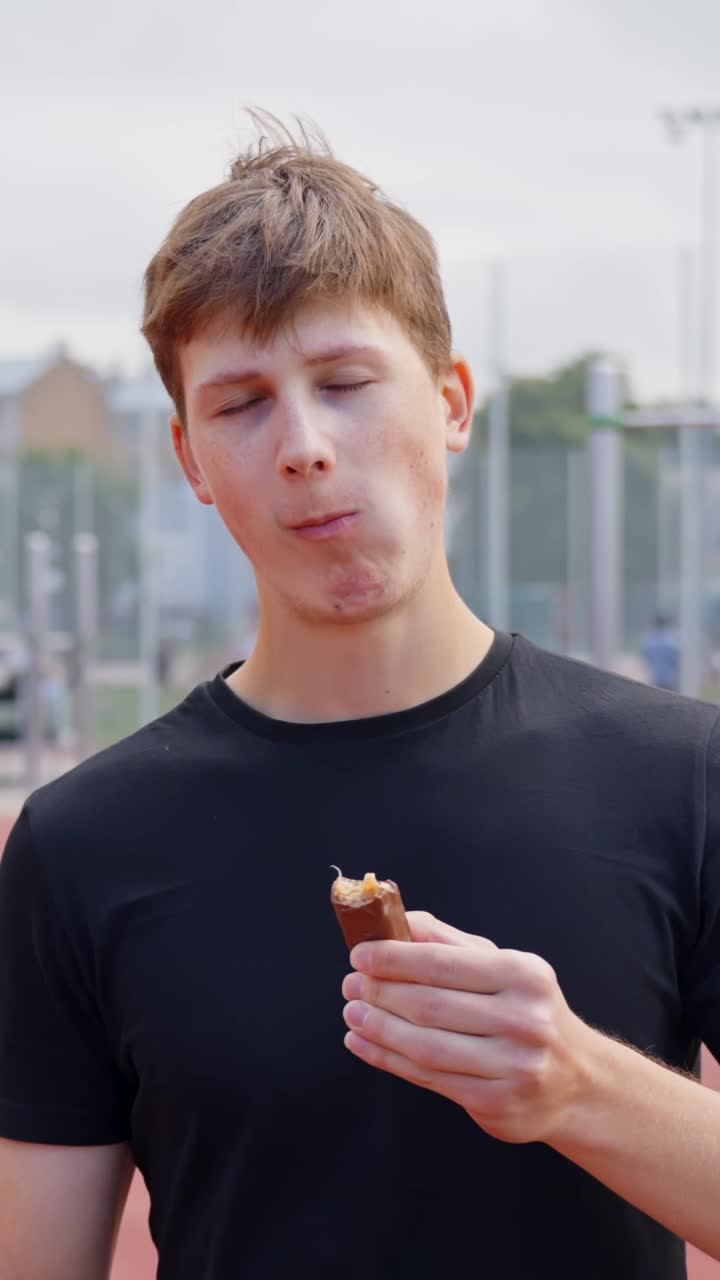 Young man smiles and takes bite of chocolate bar while relaxing after workout outdoors