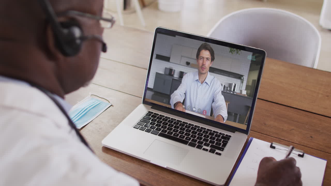 African american male doctor wearing phone headset taking notes while having a video call on laptop