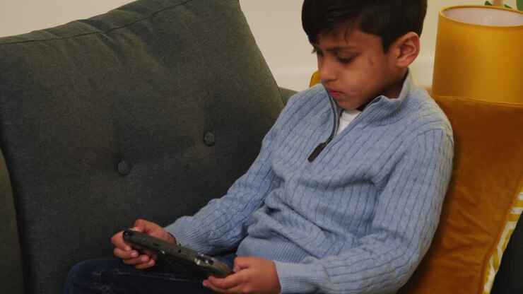 Giovane ragazzo seduto sul divano a casa che gioca o guarda contenuti in streaming su un dispositivo di gioco portatile 3