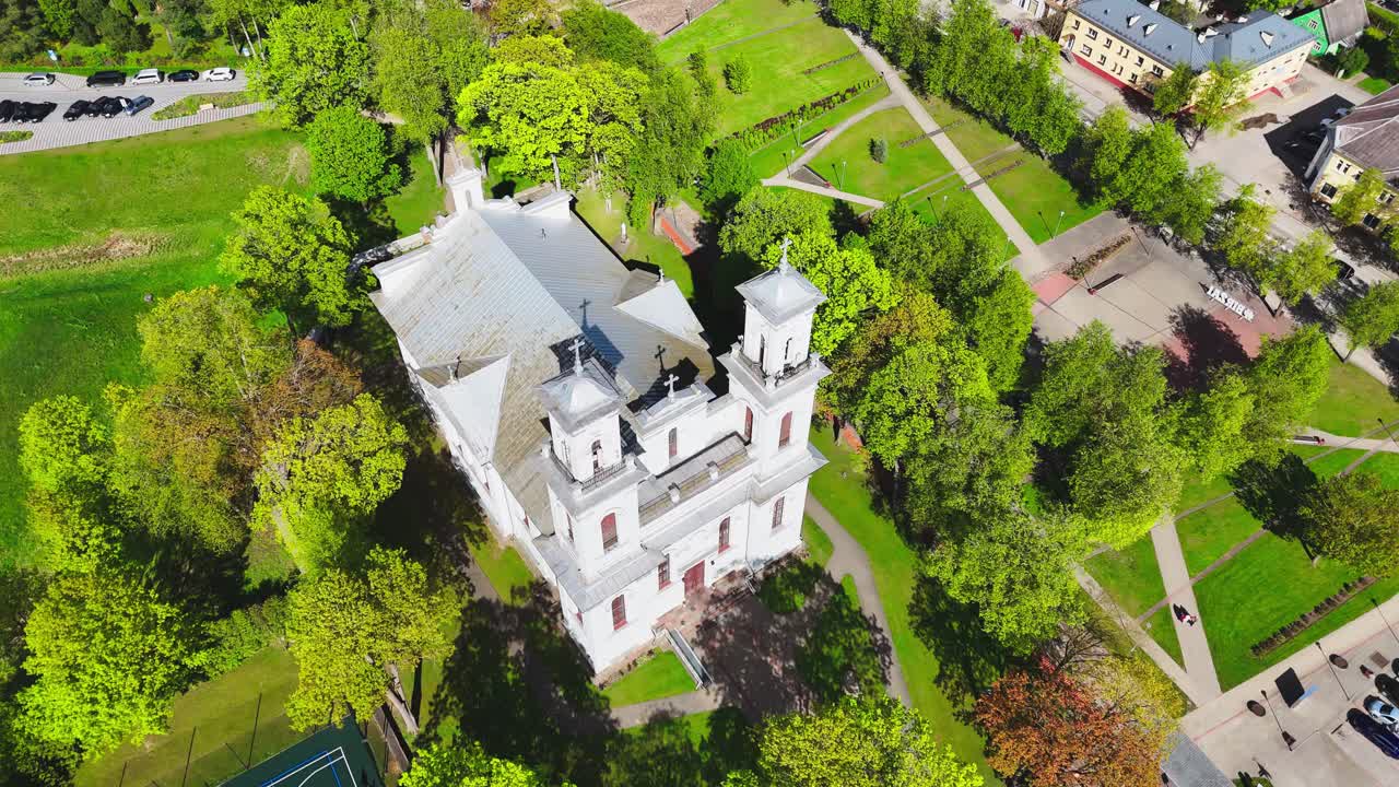 A white baroque church stands amid green trees in a Lithuanian city, surrounded by streets, buildings, and a blue basketball court on a sunny spring day.