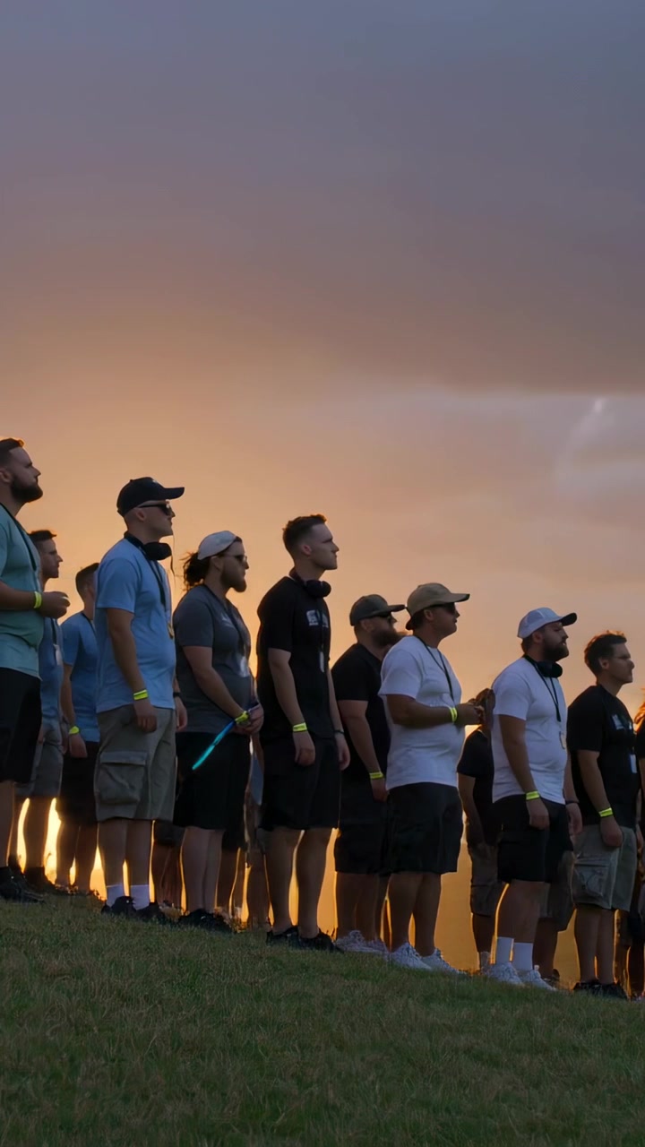 Vertical video: Watching sunset, line of men standing on slope wearing caps, lanyards, glowsticks