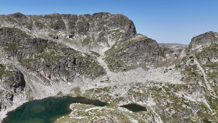 Il lago Elenino si trova sotto le ripide scogliere rocciose del picco Malyovitsa nei monti Rila sotto un cielo azzurro limpido, mostrato in una fluida ripresa aerea con drone
