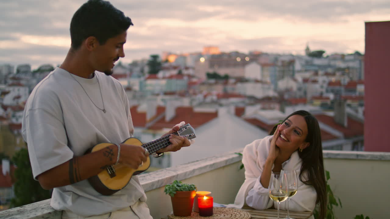 Bell'uomo che suona la chitarra su una terrazza con vista sulla città al tramonto. Appuntamento romantico di coppia
