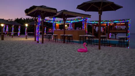 colorful string lights decorating a beach bar at dusk, creating a festive atmosphere with pink flamingo inflatable on the sand, ready for evening entertainment