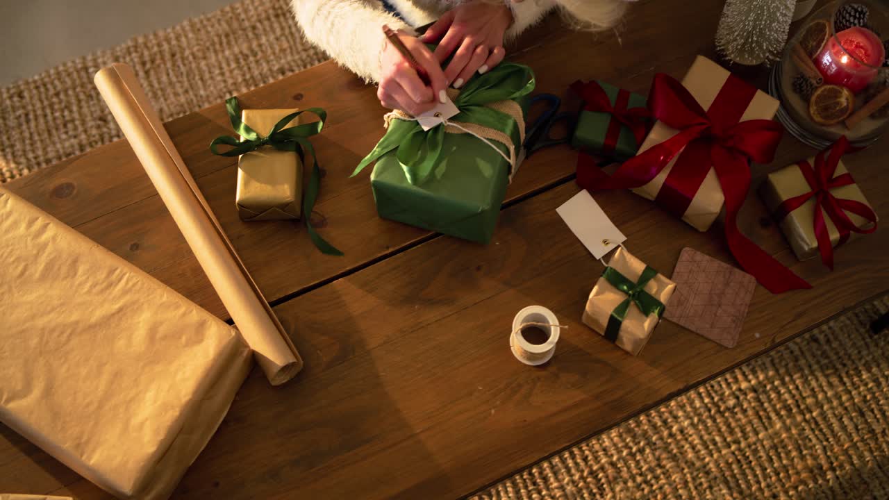 White adult woman centering green gift threading tag writing name adjusting Santa hat at home table