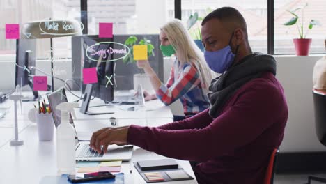 Mixed-race-businessman-wearing-face-mask-sitting-using-a-laptop-and-smartphone-in-modern-office