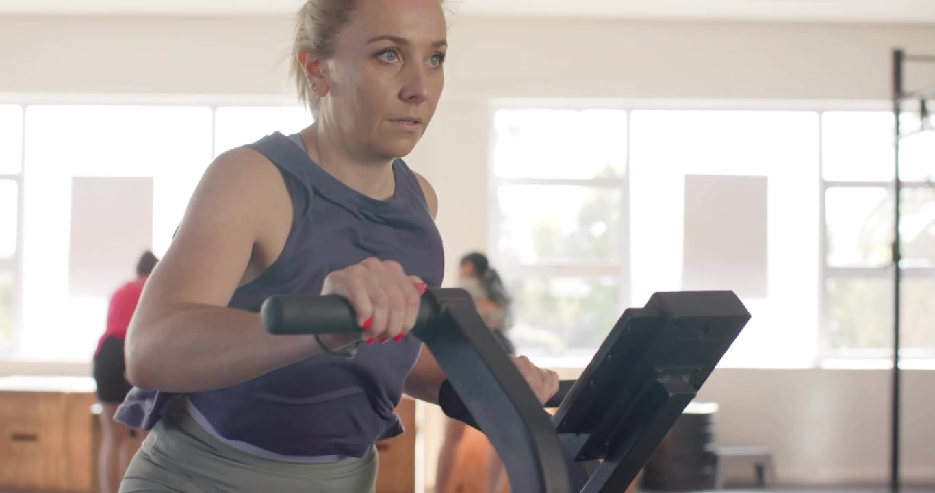La telecamera si muove verso l'alto rivelando una donna che afferra le maniglie e pedala su una cyclette a ventola in palestra, con uno scarabocchio rosa tremolante