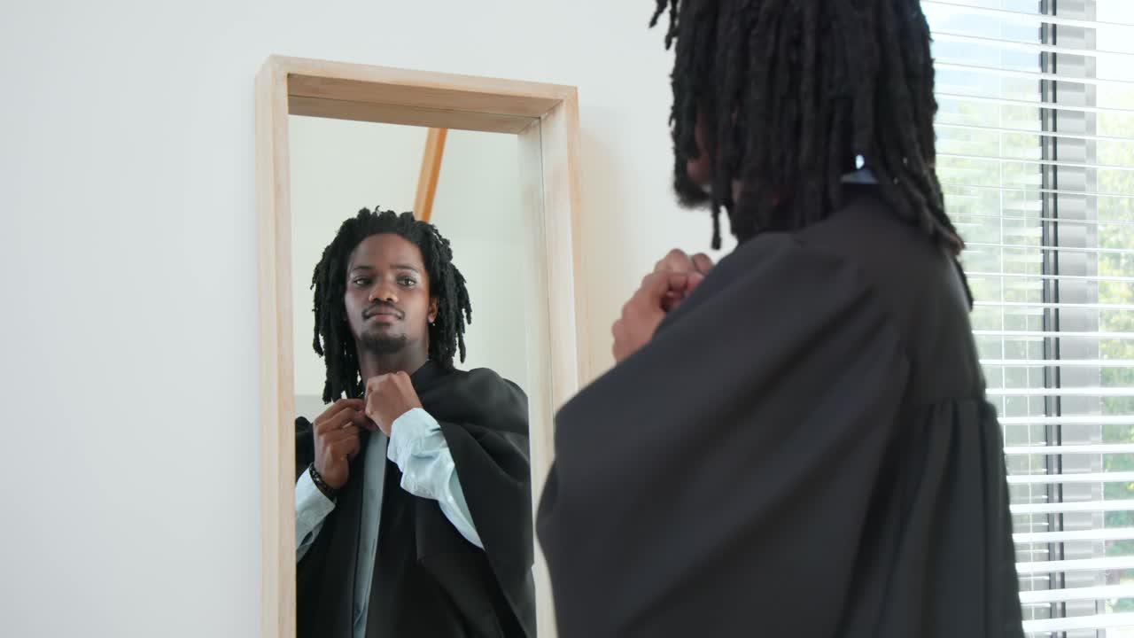 Uomo afroamericano davanti allo specchio a casa, che sistema la toga accademica nera preparandosi per la laurea.