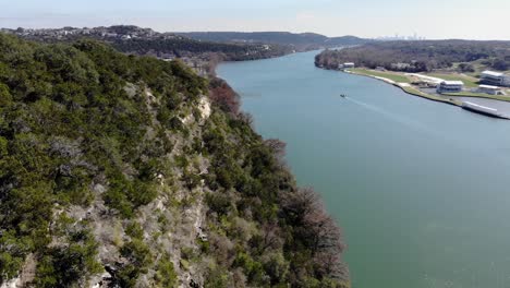 Aerial Austin Pennybacker Bridge - moving past the cliffs on the bridge overlook, moving down the river towards a small barge boat that is going towards downtown skyline in far distance.